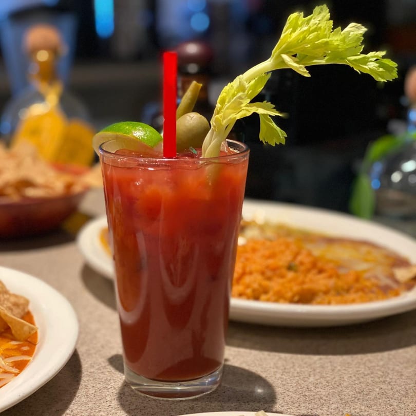 A Bloody Mary cocktail with celery, lime, and olive, next to Mexican food.
