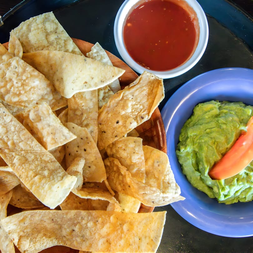 A basket of tortilla chips with a side of red salsa and guacamole with a tomato slice.